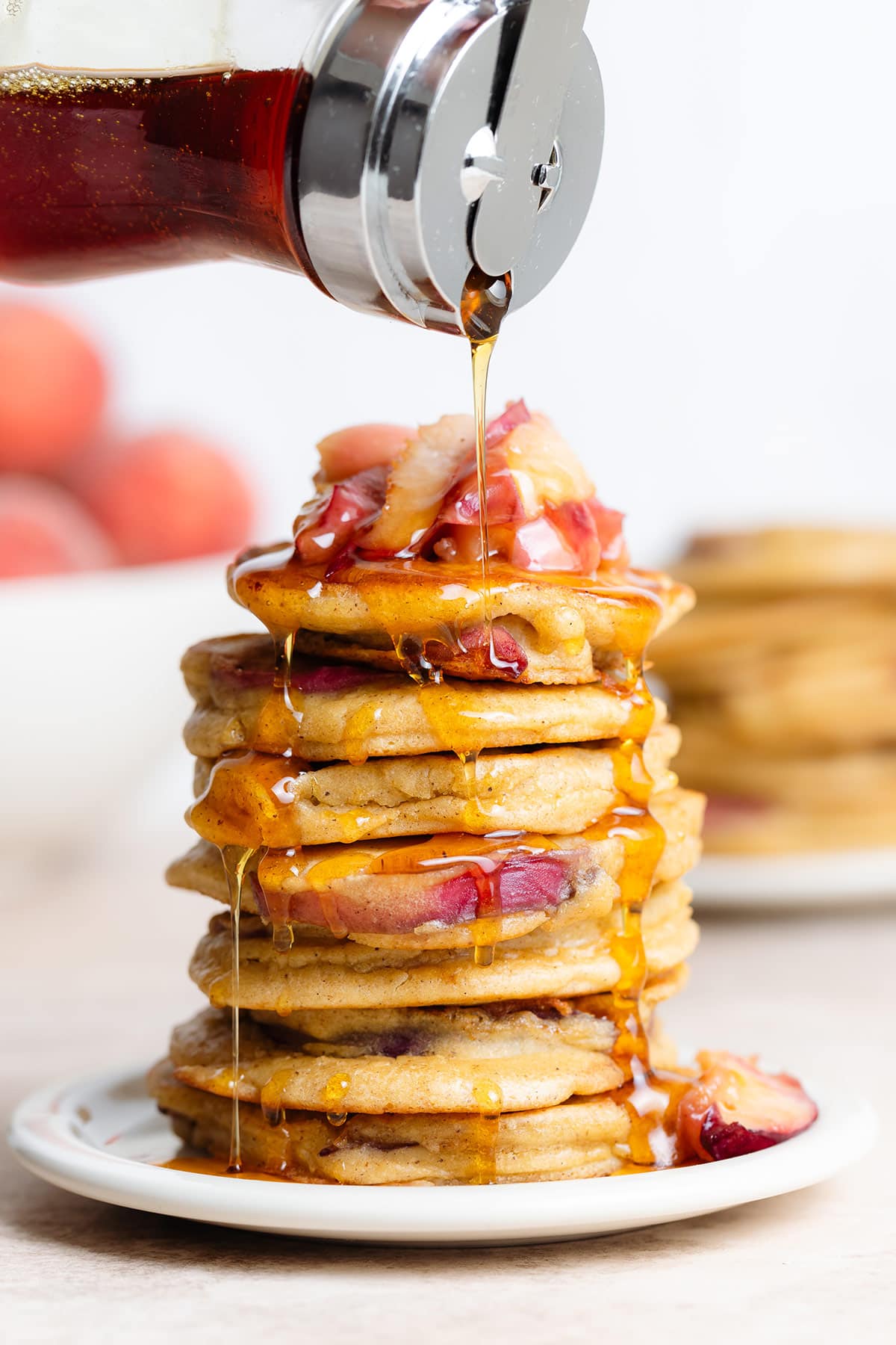 Pancakes stacked on a white plate topped with sauteed peaches and being drizzled with maple syrup.