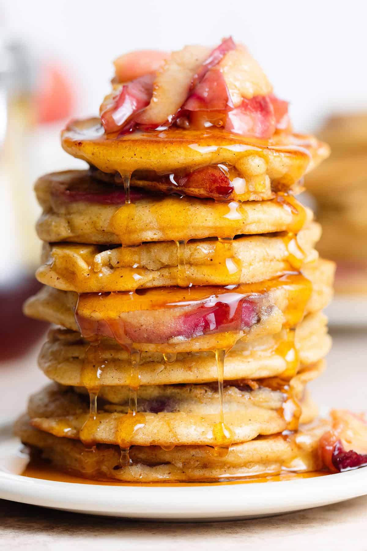 Pancakes stacked on a white plate topped with sauteed peaches and drizzled with maple syrup dripping over the edges.