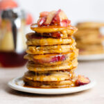 Pancakes stacked on a white plate topped with sauteed peaches and drizzled with maple syrup dripping over the edges.