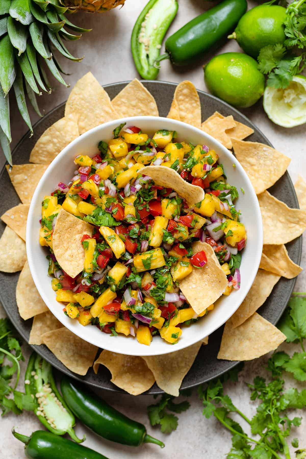 Colorful pineapple pico de gallo in a white bowl on a black plate with tortilla chips and three chips dipped in the salsa.