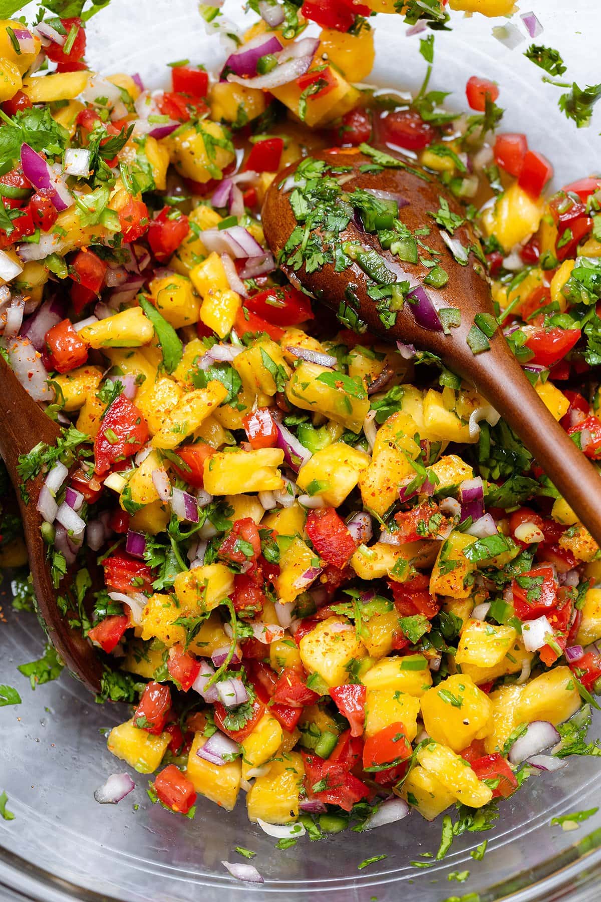 Colorful pineapple pico de gallo in a large glass bowl being mixed with two serving spoons.