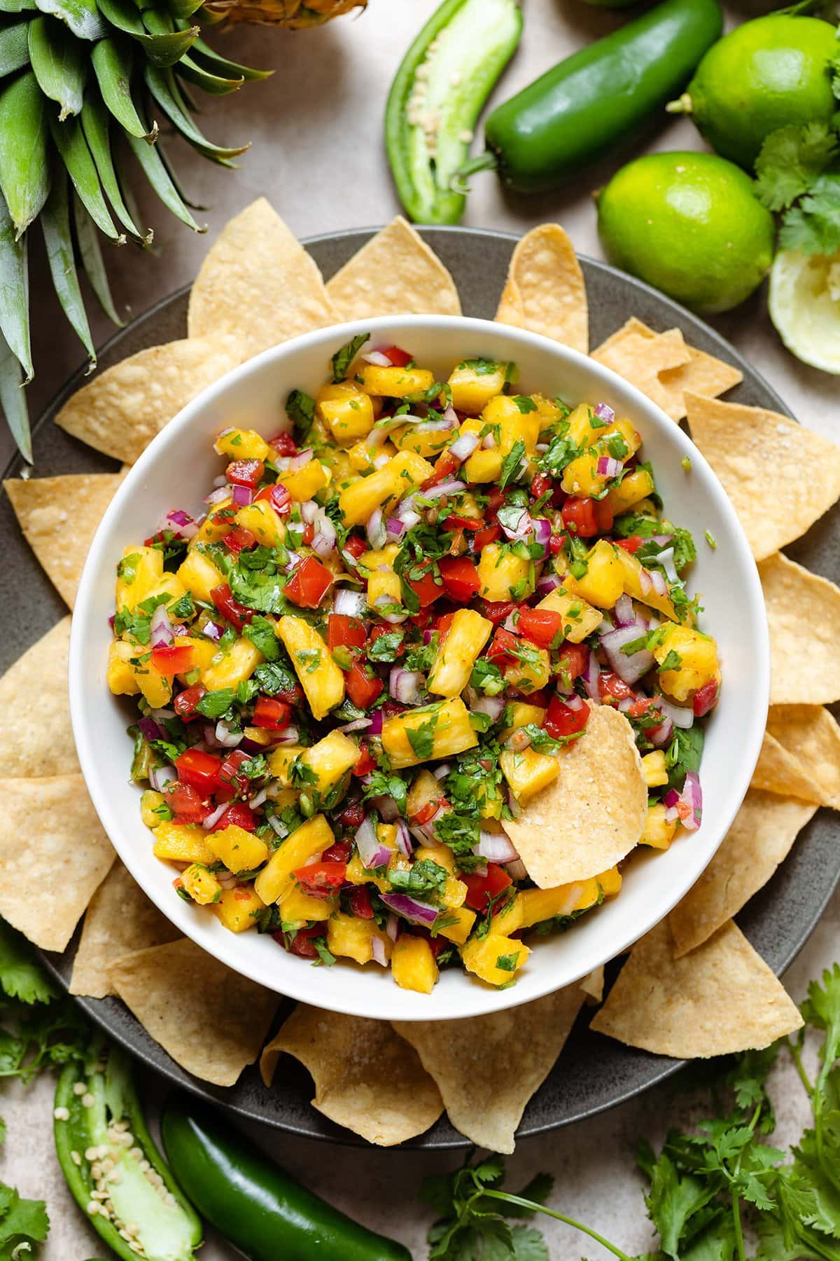 Colorful pineapple pico de gallo in a white bowl on a black plate with tortilla chips and one in the salsa.