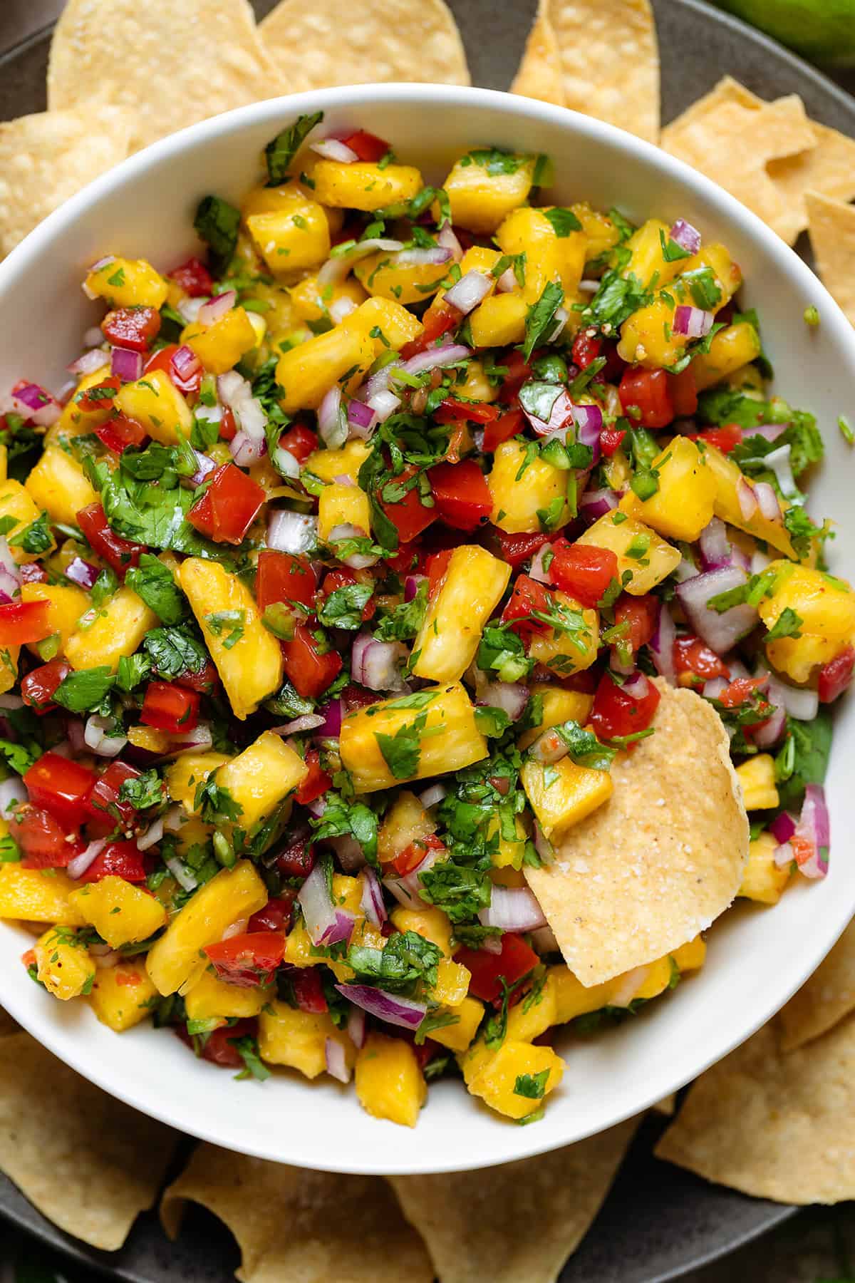 Colorful pineapple pico de gallo in a white bowl on a black plate with tortilla chips and one chip in the salsa.