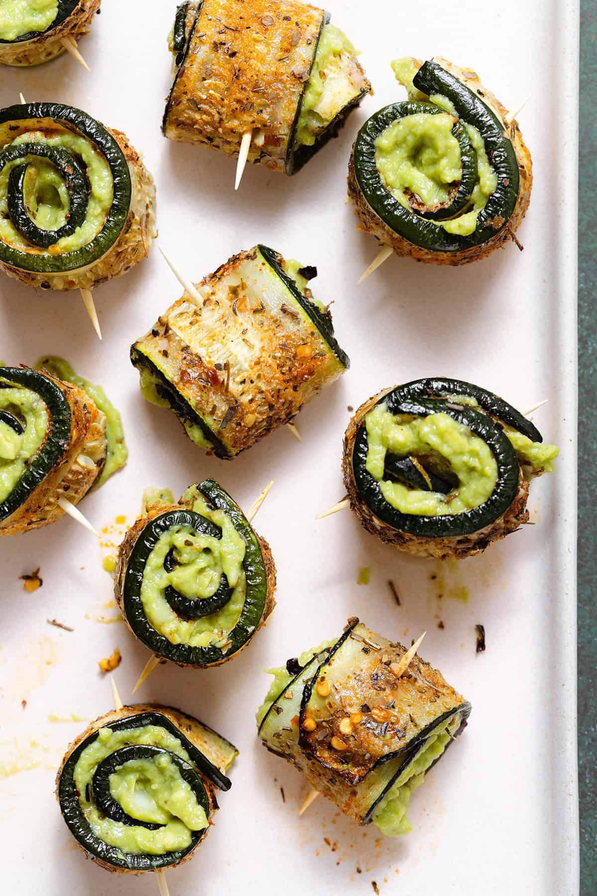 Roasted zucchini rolls with lots of spices stuffed with guacamole and secured with a toothpick on a light pink plate.