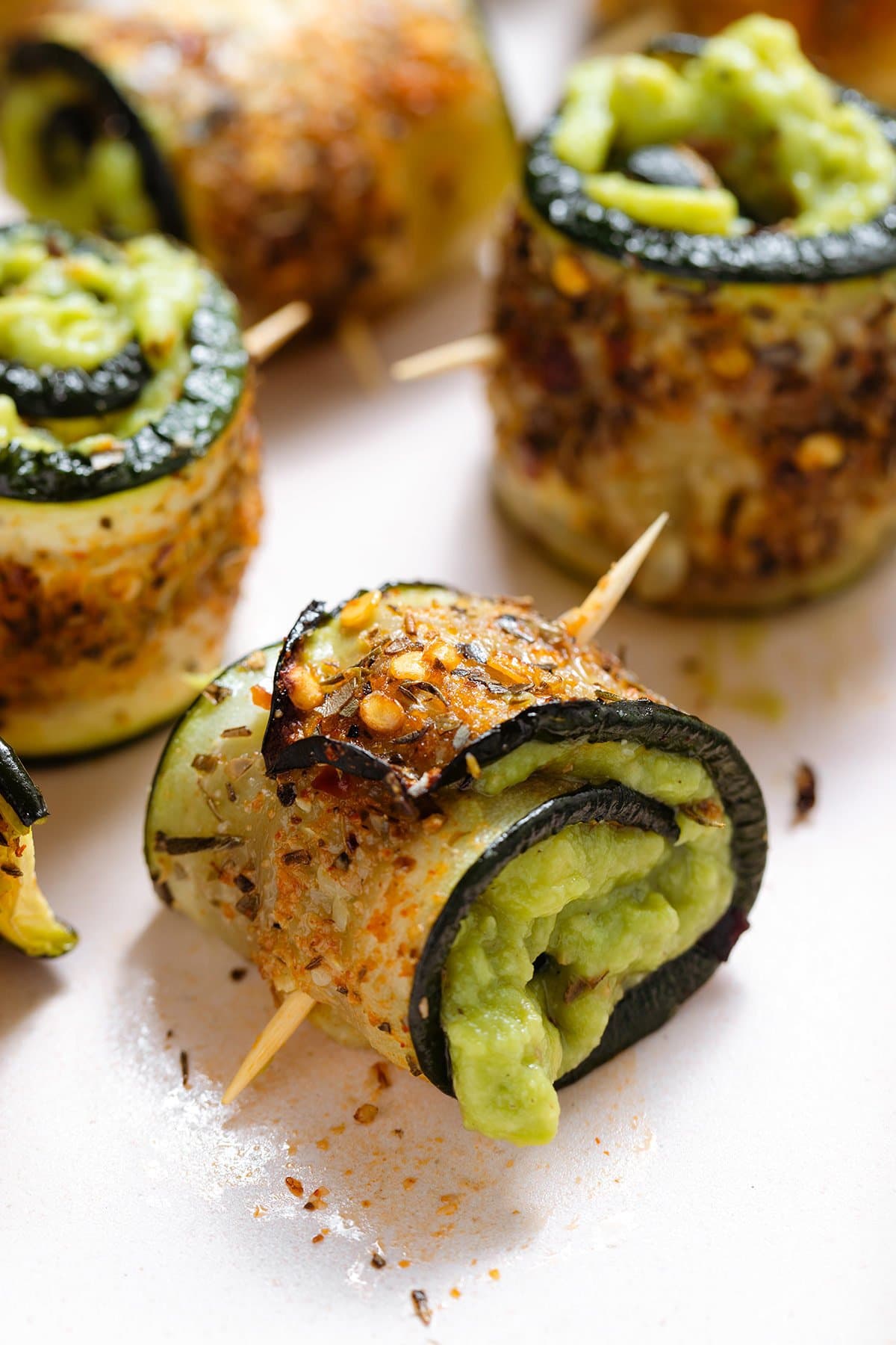 Roasted zucchini rolls with lots of spices stuffed with guacamole and secured with a toothpick on a light pink plate.