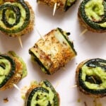 Roasted zucchini rolls with lots of spices stuffed with guacamole and secured with a toothpick on a light pink plate.