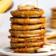 Maple syrup being poured over a stack of pancakes with banana slices on top.