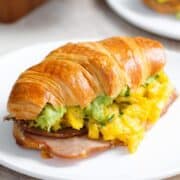 A toasted croissant filled with scrambled eggs, turkey bacon, and avocado on a white plate with more croissants in the background.