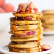 Pancakes stacked on a white plate topped with sauteed peaches and being drizzled with maple syrup.