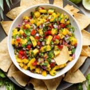Colorful pineapple pico de gallo in a white bowl on a black plate with tortilla chips and one in the salsa.