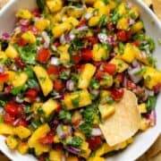 Colorful pineapple pico de gallo in a white bowl on a black plate with tortilla chips and one chip in the salsa.