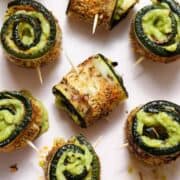 Roasted zucchini rolls with lots of spices stuffed with guacamole and secured with a toothpick on a light pink plate.