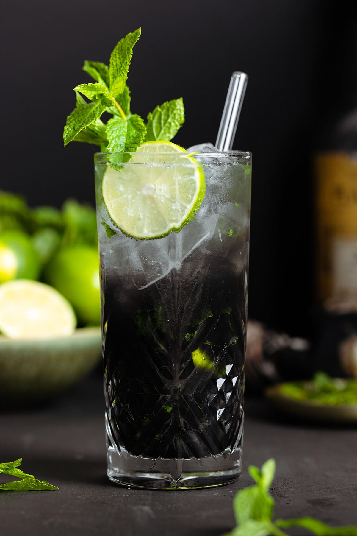 Black mojito with sparkling water on top creating an ombre effect in a tall glass garnished with a slice of lime and fresh mint.
