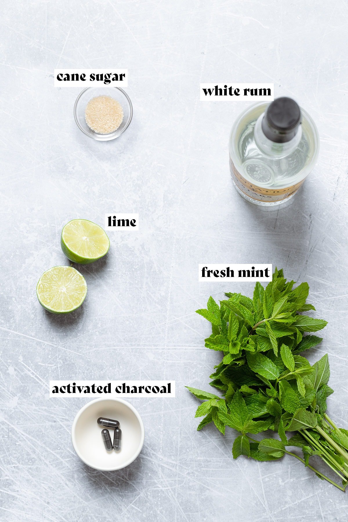 Ingredients for black mojito like rum, mint, and charcoal capsuled on a grey background.