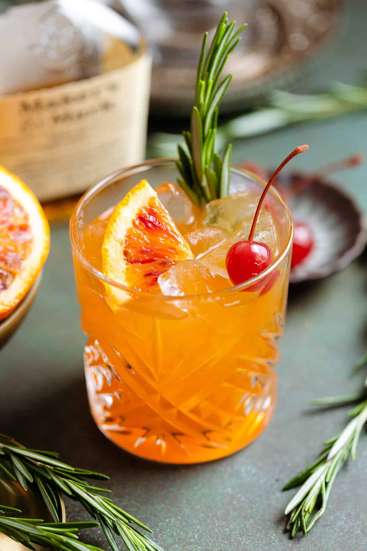 Orange old fashioned in a short glass garnished with a cherry, fresh rosemary spring, and a wedge of a blood orange.