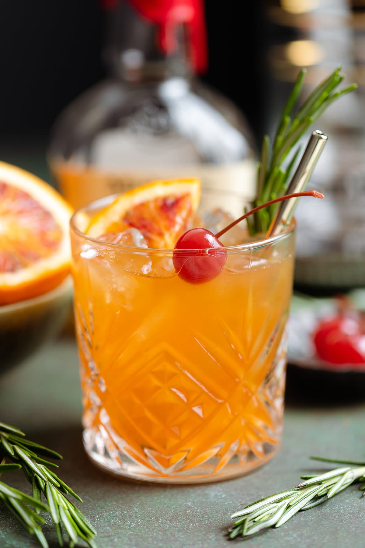 Orange old fashioned in a short glass garnished with a cherry, fresh rosemary spring, and a wedge of a blood orange.