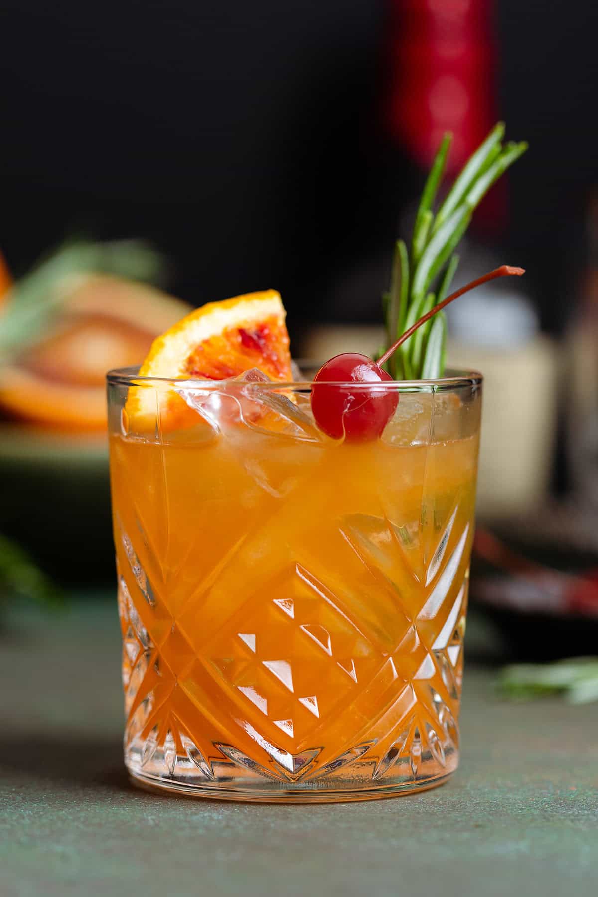 Orange old fashioned in a short glass garnished with a cherry, fresh rosemary spring, and a wedge of a blood orange.