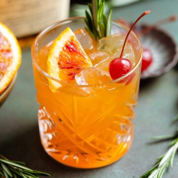Orange old fashioned in a short glass garnished with a cherry, fresh rosemary spring, and a wedge of a blood orange.