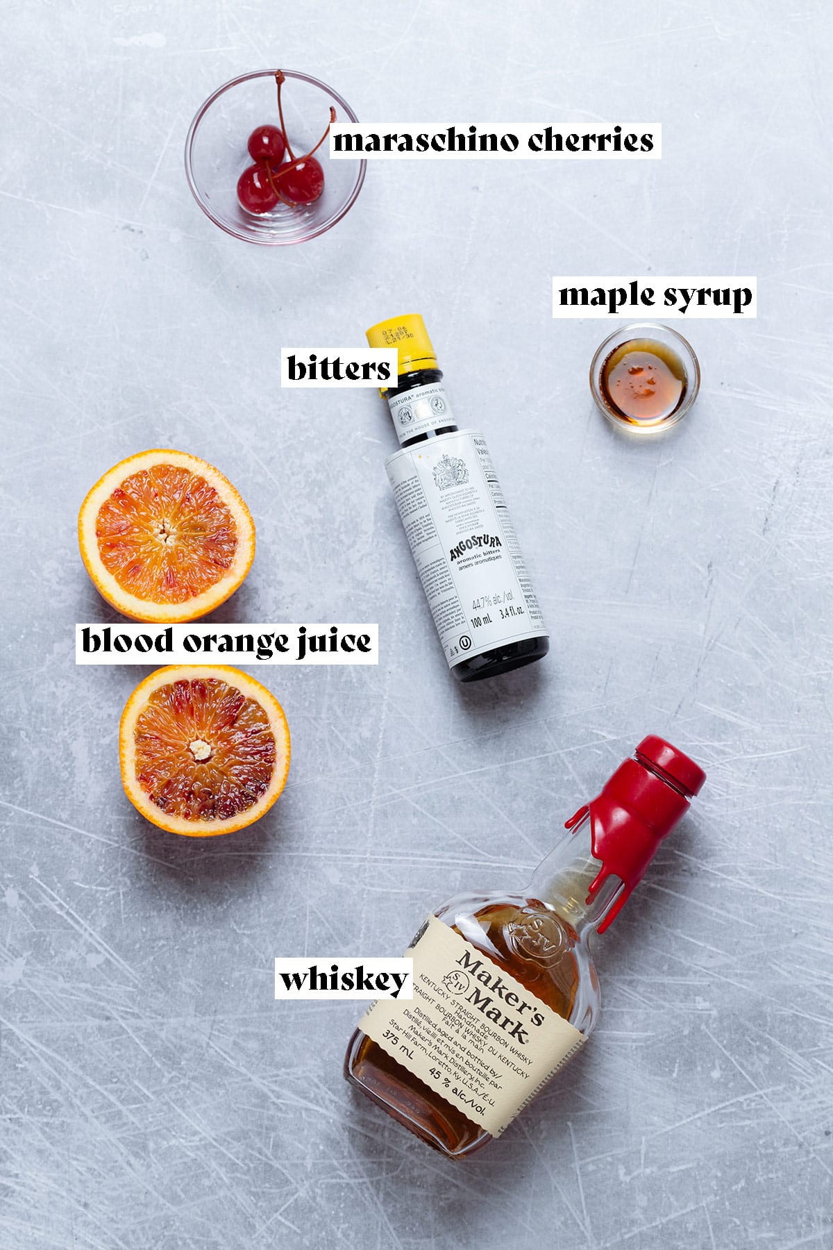 Ingredients for an orange old fashioned like whiskey, bitters, and fresh orange laid out on a grey background.