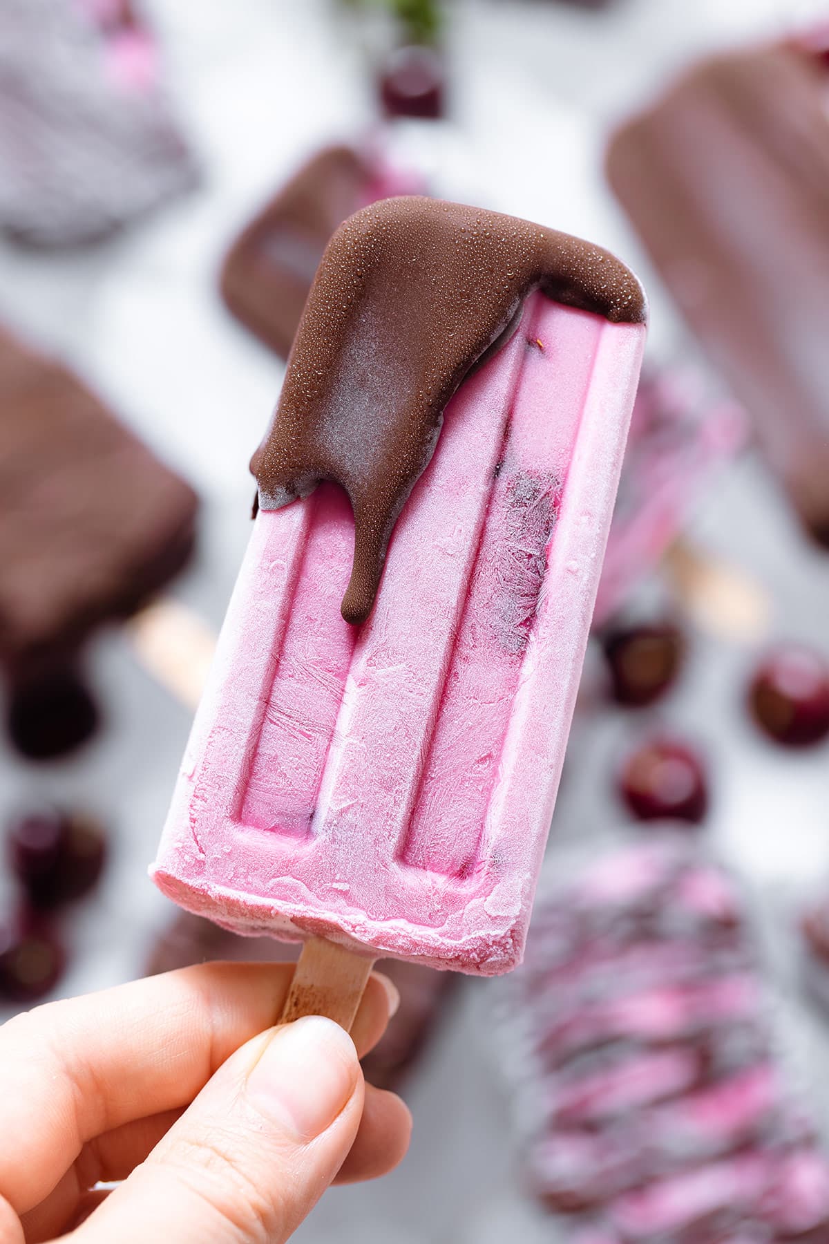 A hand holding a pink popsicle partially covered with chocolate at the tip.