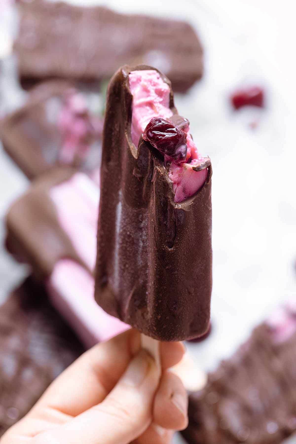 A hand holding a pink popsicle completely covered with chocolate with the corner bitten off.