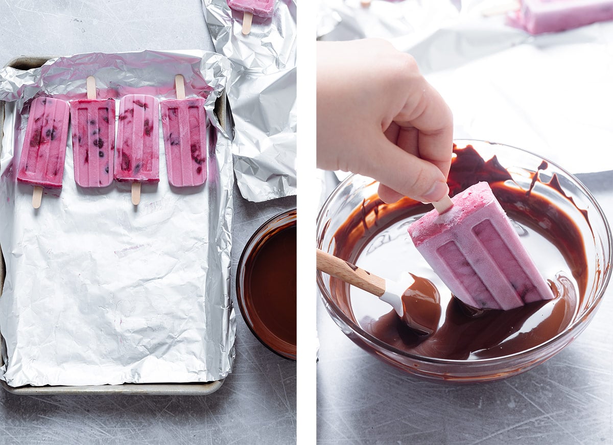 Pink cherry popsicles on a baking sheet being dipped into melted chocolate one by one.