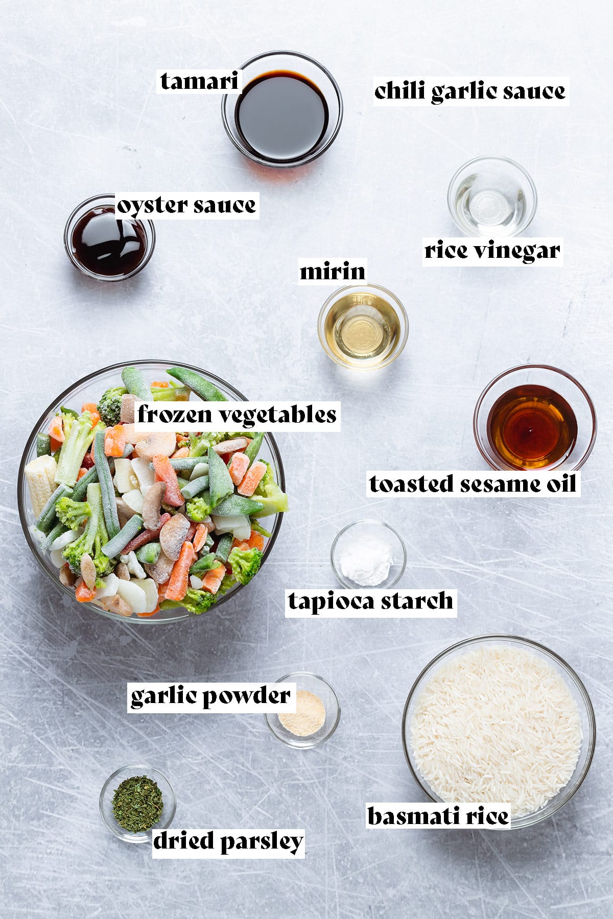 Ingredients for a stir fry like frozen vegetables, rice, and condiments laid out on a grey background.