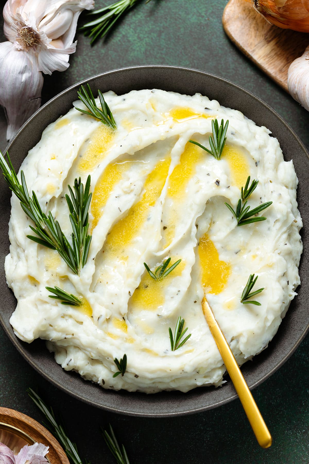 Mashed potatoes with fresh rosemary and melted butter in a black bowl with a gold spoon on the right side.