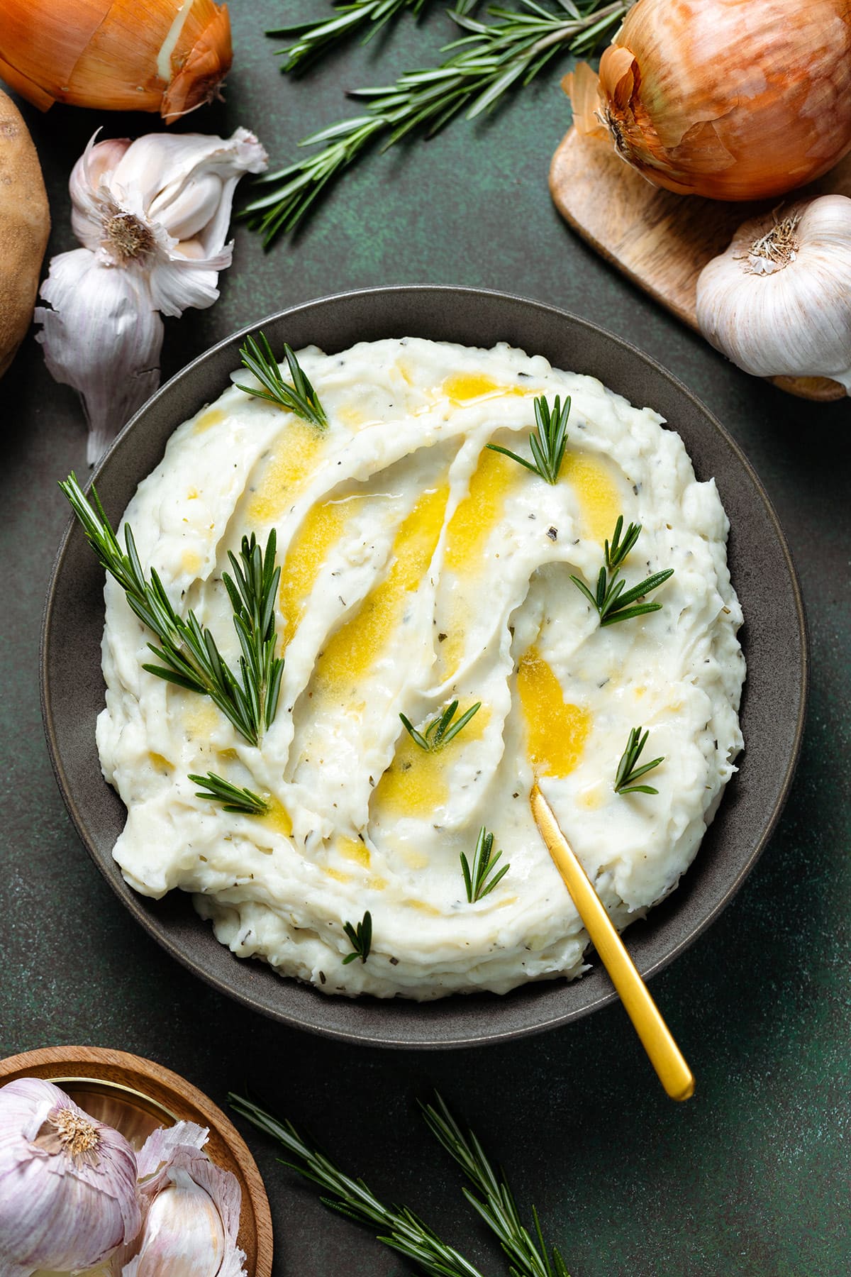 Mashed potatoes with fresh rosemary and melted butter in a black bowl with a gold spoon on the right side.