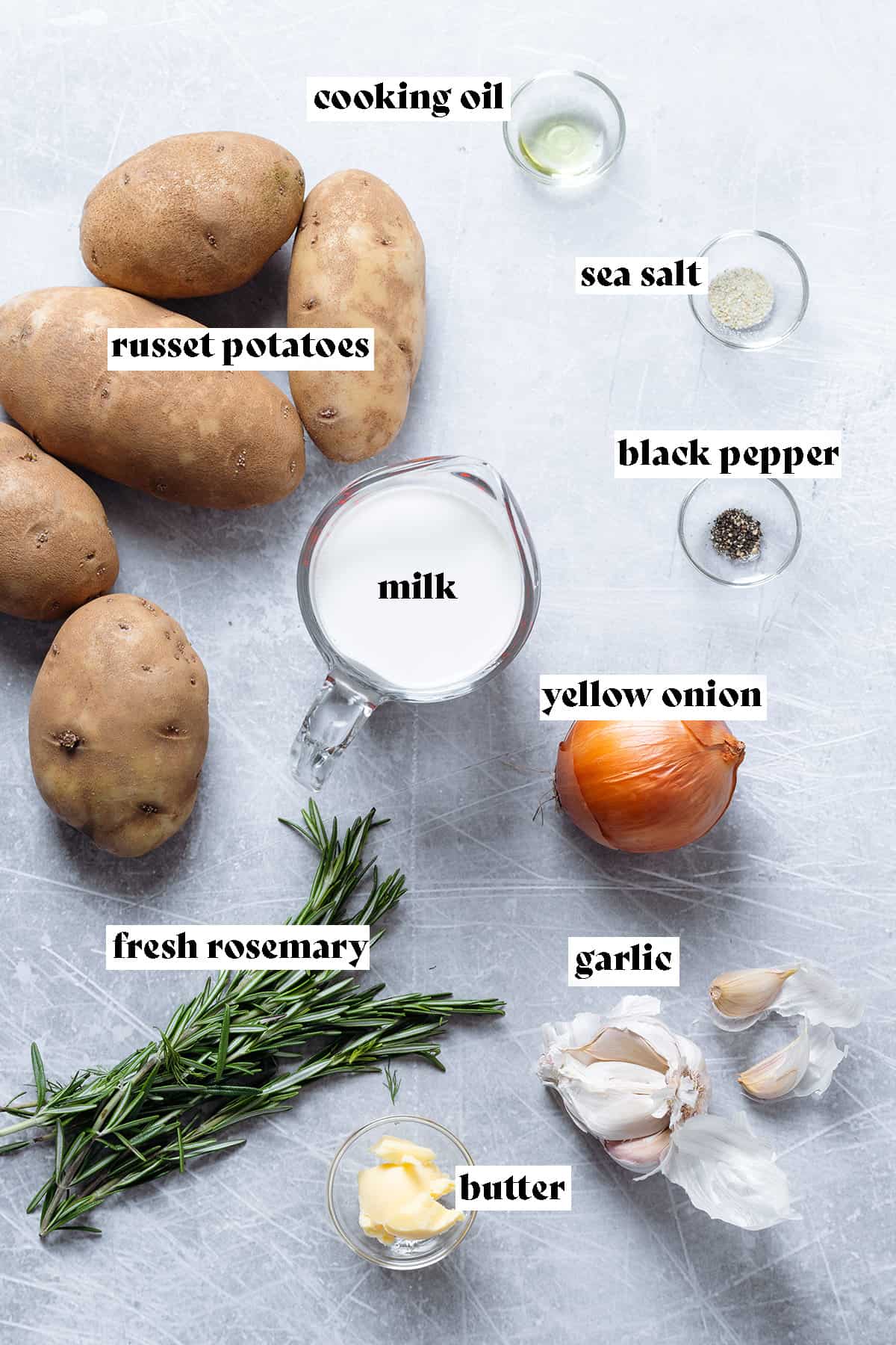 Ingredients for mashed potatoes like milk, potatoes, fresh rosemary, and onion on a grey background.