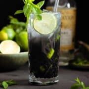 Black mojito with sparkling water on top creating an ombre effect in a tall glass garnished with a slice of lime and fresh mint.