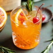 Orange old fashioned in a short glass garnished with a cherry, fresh rosemary spring, and a wedge of a blood orange.