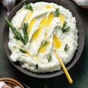 Mashed potatoes with fresh rosemary and melted butter in a black bowl with a gold spoon on the right side.