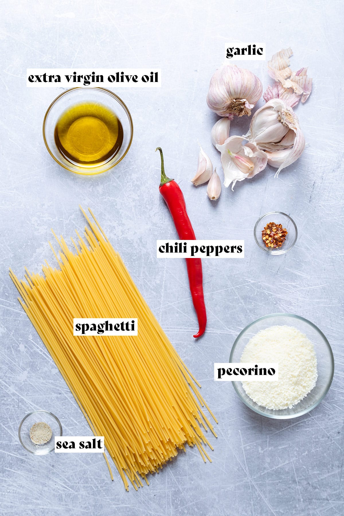 Ingredients like spaghetti, olive oil, garlic, and chili pepper all laid out on a grey background.