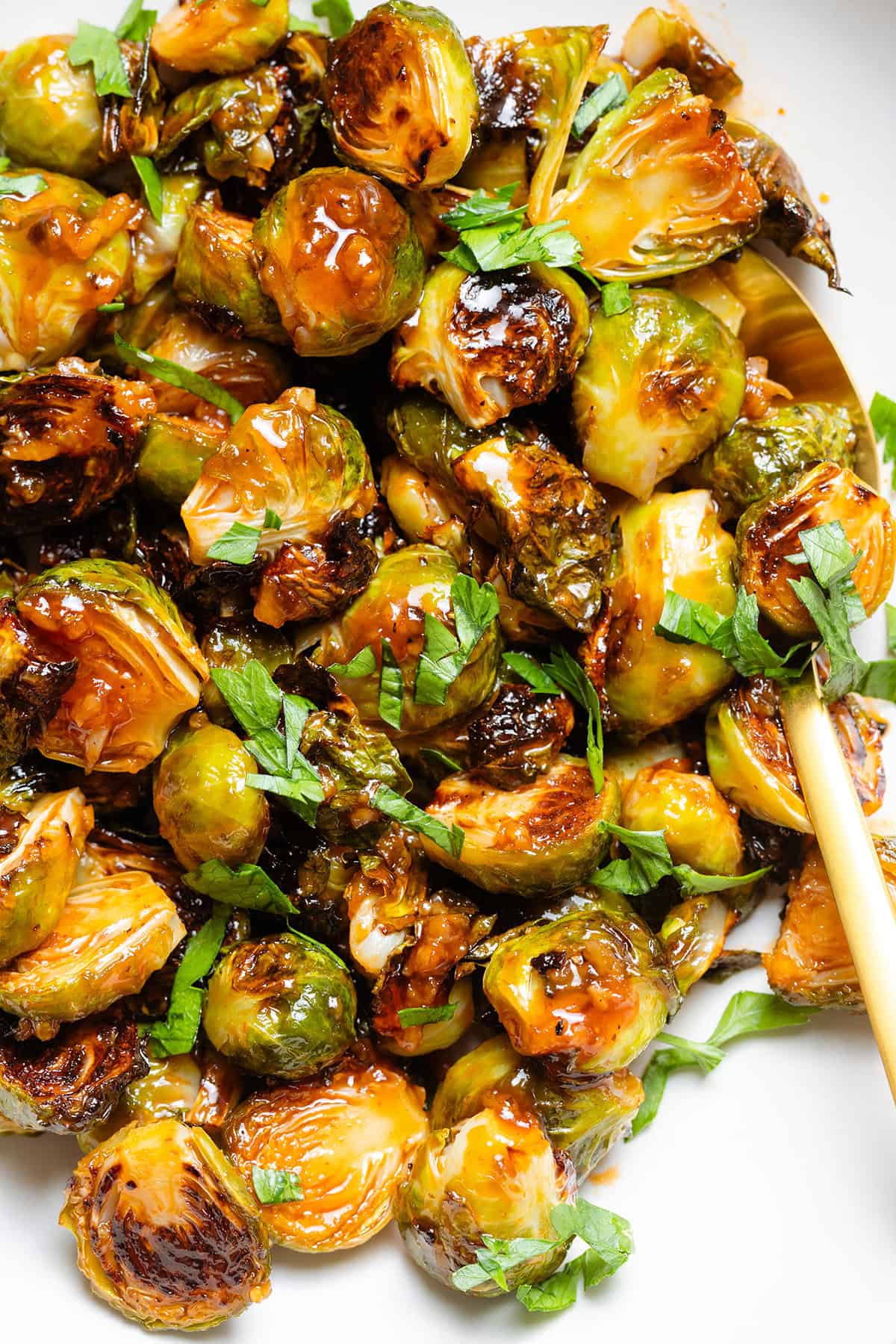 Charred saucy brussels sprouts in a white bowl with a gold serving spoon on the right.