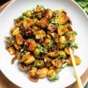 Charred saucy brussels sprouts in a white bowl with a gold serving spoon on the right.