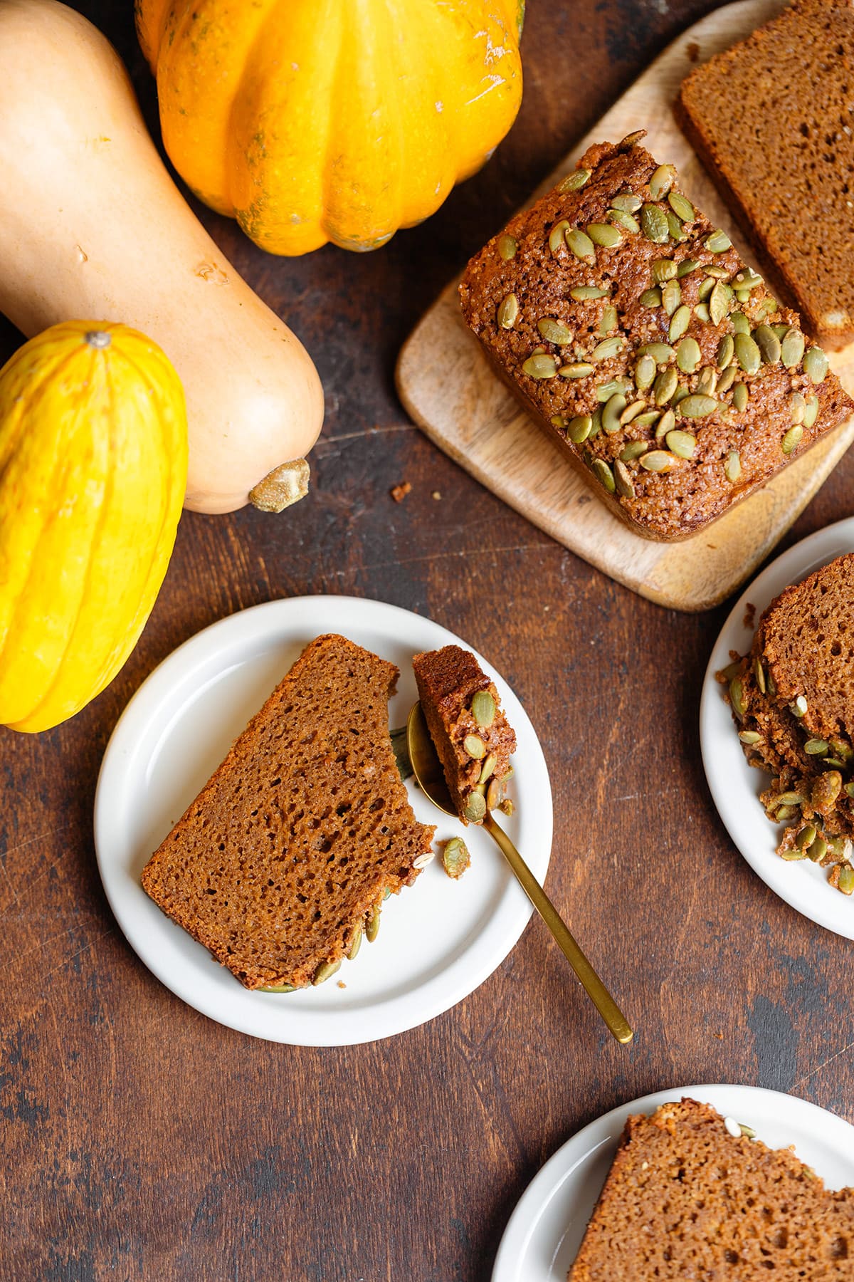 Sliced pumpkin bread on small white plates with a spoon cutting off a corner of one slice. Pumpkins on the top left of the frame.