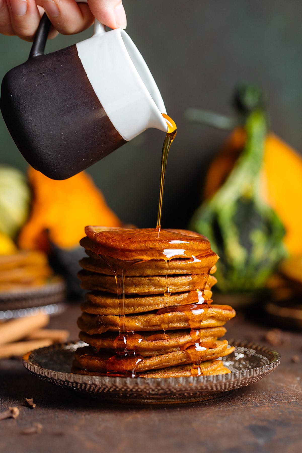 Pumpkin pancakes stacked on a brown glass plate with maple syrup being poured over the top.