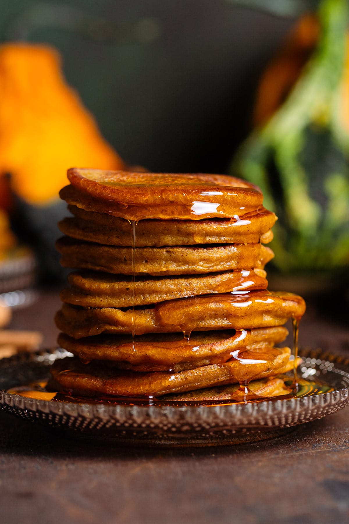 Pumpkin pancakes stacked on a brown glass plate with maple syrup dripping down the sides, with various pumpkins in the background.