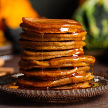 Pumpkin pancakes stacked on a brown glass plate with maple syrup dripping down the sides, with various pumpkins in the background.