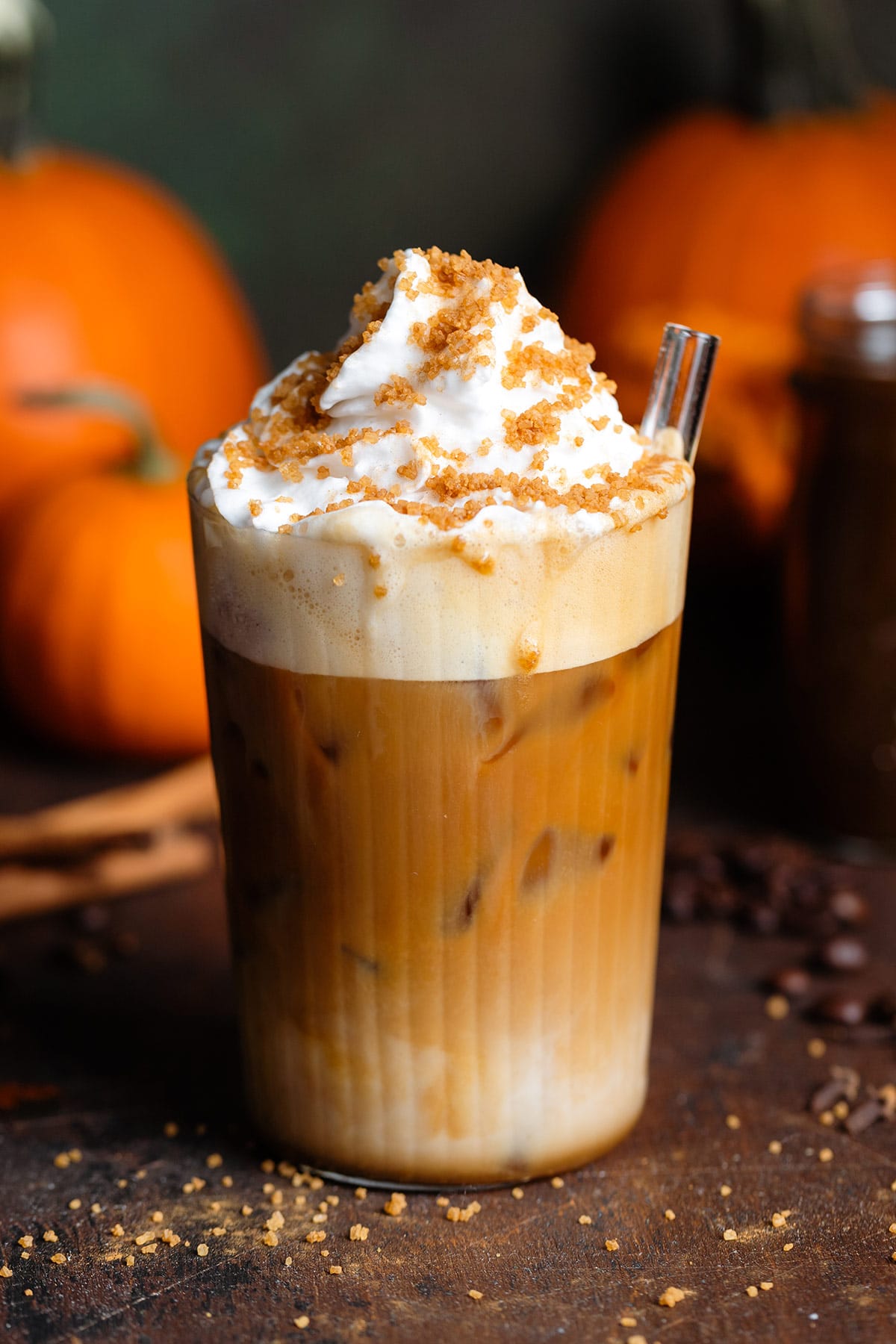 Iced pumpkin latte with whipped cream and sugar topping in a tall glass on a wooden backdrop with pumpkins in the background.