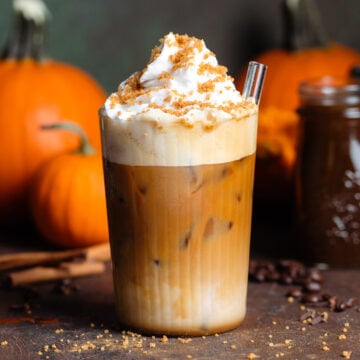 Iced pumpkin latte with whipped cream and sugar topping in a tall glass on a wooden backdrop with pumpkins in the background.