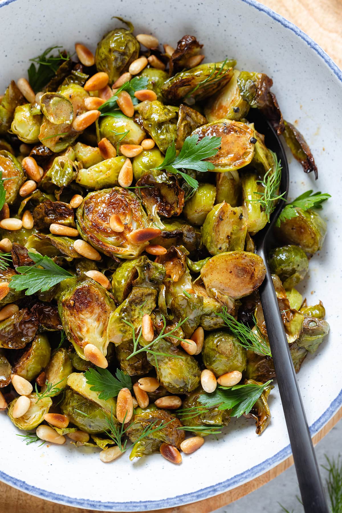 Charred brussels sprouts in a white bowl topped with fresh parsley, and toasted pine nuts with a spoon inserted on the right.
