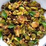 Charred brussels sprouts in a white bowl topped with a mustard vinaigrette, fresh parsley, and toasted pine nuts.