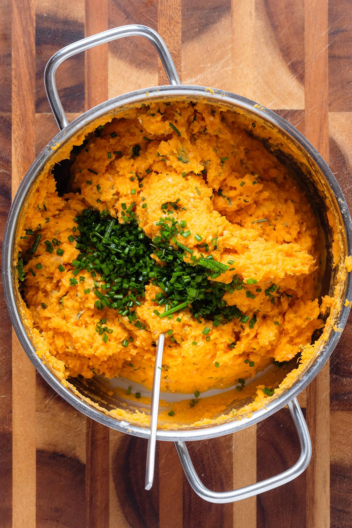 Mashed sweet potatoes in a large pot with fresh chopped chives and a large spoon.