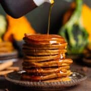 Pumpkin pancakes stacked on a brown glass plate with maple syrup being poured over the top.
