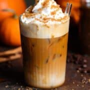 Iced pumpkin latte with whipped cream and sugar topping in a tall glass on a wooden backdrop with pumpkins in the background.