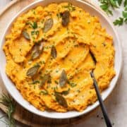 Mashed sweet potatoes in a white bowl topped with fried sage leaves and chives.