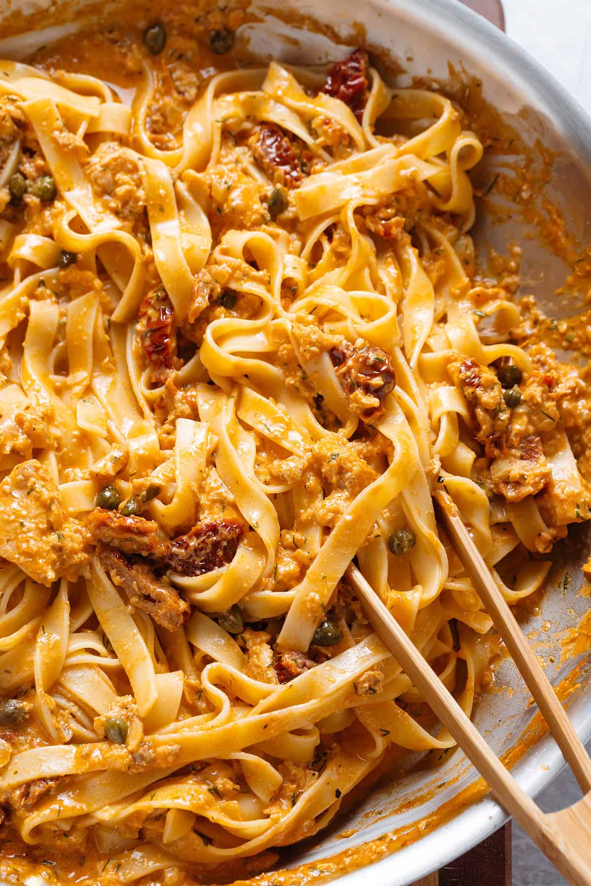 Fettuccine with creamy sundried tomato sauce and canned salmon in large skillet with wooden tongs on the right.