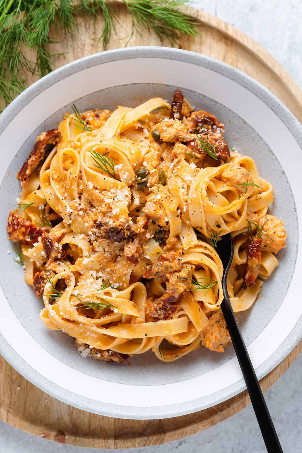 Fettuccine with creamy sundried tomato sauce and canned salmon in a low grey bowl topped with fresh dill and parmesan with a black fork on the right.